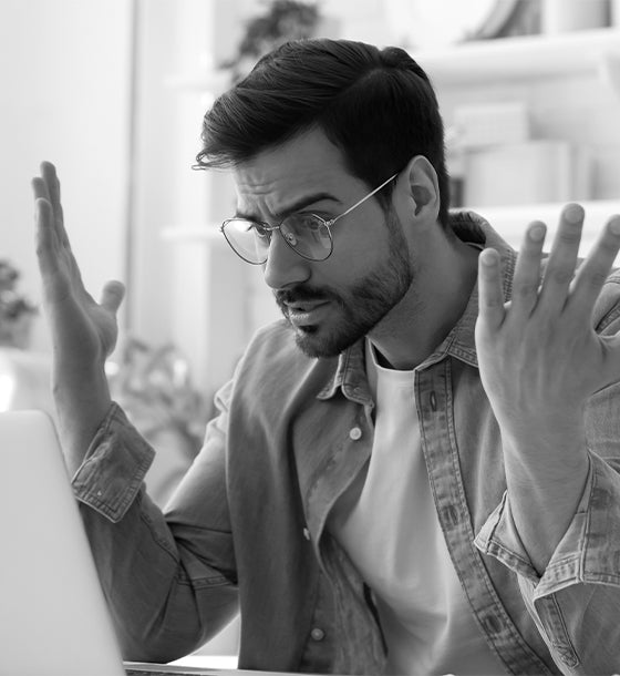Homme portant des lunettes et une chemise en jean, levant les mains avec une expression d’incompréhension en regardant un écran d'ordinateur.